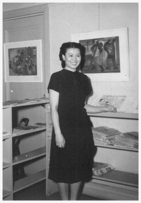 Black and white photograph of an Asian American woman posing in front of small drawings and paintings.